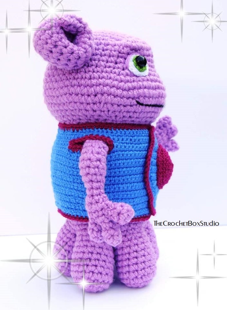 May include: A crocheted purple plush toy with a blue vest and a red heart. The toy has a large head with a green eye and a small mouth.