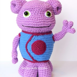 May include: A crocheted purple alien plush toy with a blue vest and a red circle on the front. The toy has large green eyes and a friendly expression. The toy is standing on a white surface.