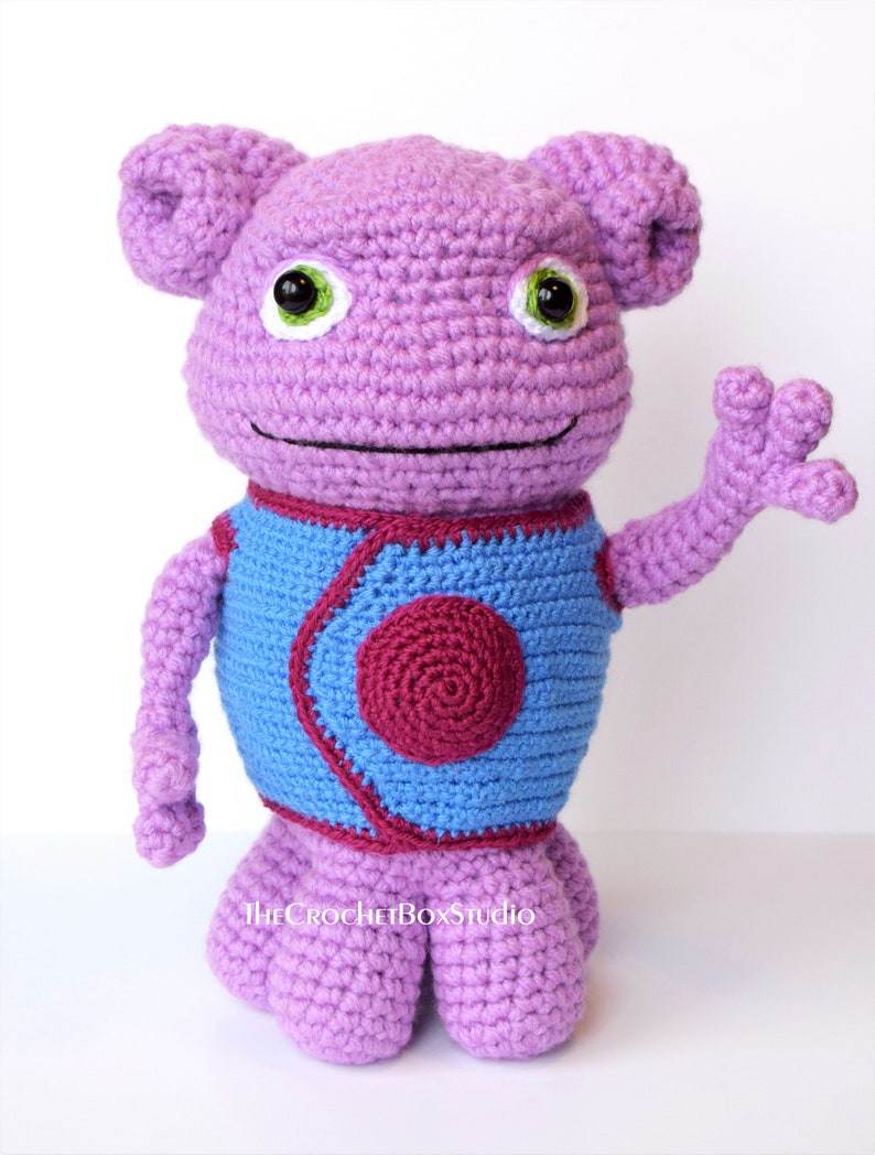 May include: A crocheted purple alien plush toy with a blue vest and a red circle on the front. The toy has large green eyes and a friendly expression. The toy is standing on a white surface.