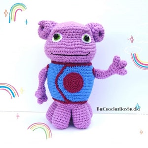 May include: A crocheted purple alien toy with green eyes and a blue and red vest. The toy is standing on a white background with rainbow drawings.