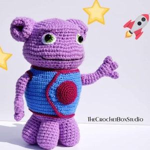 May include: Purple crocheted alien toy with blue and red details, standing on a white background. The alien has large green eyes and a friendly expression. The toy is holding out its right arm, as if waving. The text "THECROCHETBOXStudio" is visible in the lower right corner of the image.