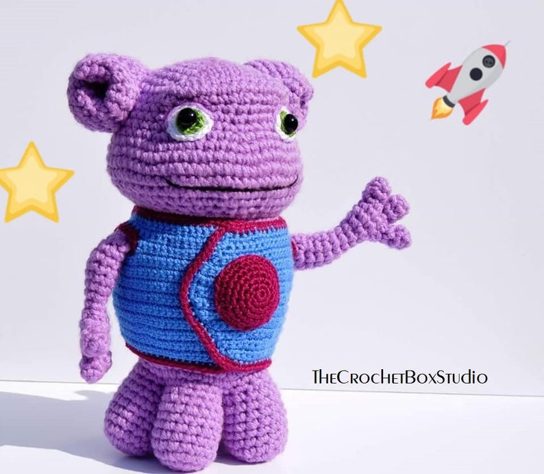 May include: Purple crocheted alien toy with blue and red details, standing on a white background. The alien has large green eyes and a friendly expression. The toy is holding out its right arm, as if waving. The text "THECROCHETBOXStudio" is visible in the lower right corner of the image.