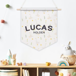May include: A white banner with a pattern of yellow flowers and white geese. The banner has the name "LUCAS HOLDEN" printed in black letters.