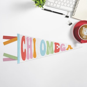 May include: White pennant flag with the text "CHI OMEGA" in colorful, retro-style lettering. The pennant has a light blue border and colorful felt streamers. The pennant is on a white surface with office supplies and a cup of coffee.