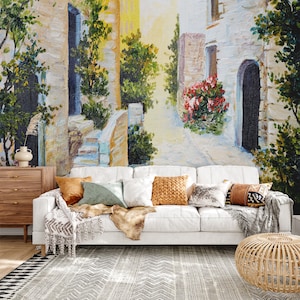 May include: A white couch with pillows and a throw blanket sits in front of a wall mural depicting a colorful, sunny, European street scene with stone buildings, flowers, and trees.