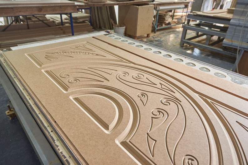 May include: A large, unfinished wooden door with intricate carved floral and scrollwork designs. The door is light brown and sits on a manufacturing machine, showcasing the detailed craftsmanship. The image is taken in a workshop setting.