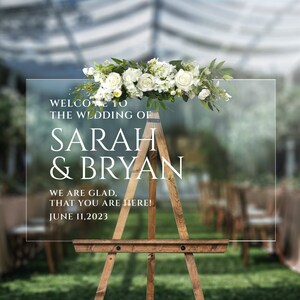 May include: A clear acrylic wedding sign with white text on a wooden easel. The sign reads "Welcome to the wedding of Sarah & Bryan, We are glad that you are here! June 11, 2023". The sign is decorated with white flowers.