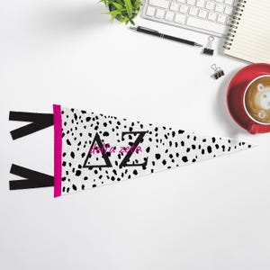 Könnte beinhalten: Ein weißer Wimpel mit einem schwarz-weißen Dalmatiner-Druck. Der Wimpel hat einen rosa Rand und schwarze Bänder. Die Buchstaben "AZ" sind in Schwarz mit den Worten "delta zeta" in rosa Schrift.