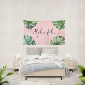 Könnte beinhalten: Ein rosa Wandteppich mit grünen tropischen Blättern und dem Text "Alpha Phi" in schwarzer Schrift.