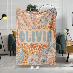 May include: A soft, plush throw blanket with a retro floral pattern in shades of blue, orange, and yellow. The blanket features the name "Olivia" in a large, blue font.