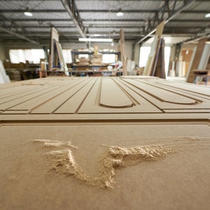 May include: Close-up of a large, unfinished wooden door panel with intricate carved designs. The panel is light brown and shows the detail of the woodworking process, with visible grooves and cutouts. The workshop background is blurred.
