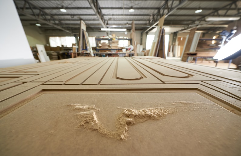 May include: Close-up of a large, unfinished wooden door panel with intricate carved designs. The panel is light brown and shows the detail of the woodworking process, with visible grooves and cutouts. The workshop background is blurred.