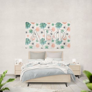 Könnte beinhalten: Ein weißer Wandteppich mit einem floralen Muster aus Sukkulenten und rosa Blüten. Die Buchstaben "AEΦ" befinden sich in der Mitte des Wandteppichs.