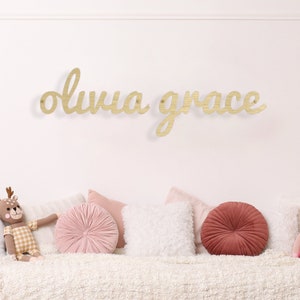 May include: A wooden sign with the name "Olivia Grace" written in cursive script. The sign is hanging on a white wall above a white couch with pink and red pillows.