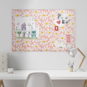 May include: A rectangular bulletin board with a daisy pattern in white and yellow on a pink background. The board displays children's drawings, photos, and a red letter "G" magnet. A desk lamp and desk are in the foreground.
