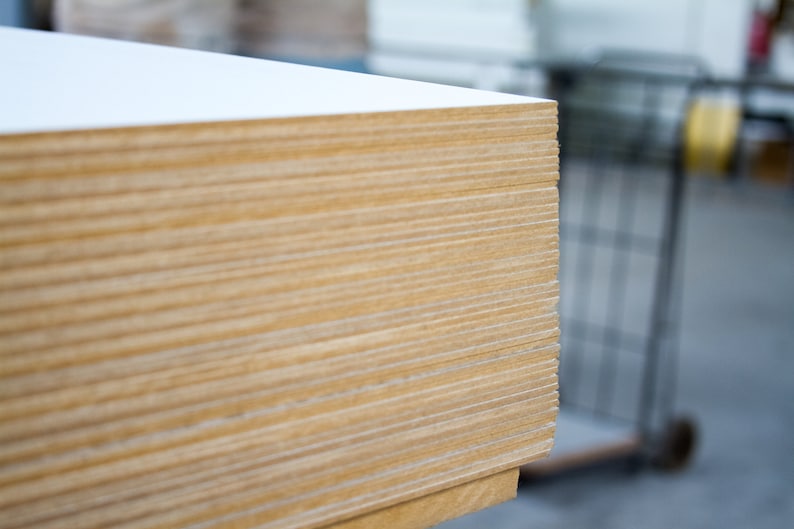 May include: A stack of rectangular wooden boards with a white top layer. The edges of the boards reveal a layered, light brown wood grain. The boards are likely for construction or woodworking projects.