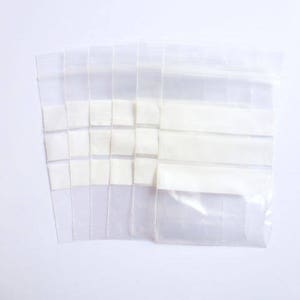 Könnte beinhalten: Mehrere transparente, wiederverschließbare Plastiktüten liegen auf einer weißen Oberfläche. Die Beutel sind rechteckig und haben einen Reißverschluss. Einige Beutel sind mit weißen, rechteckigen Objekten gefüllt. Die Beutel sind transparent.