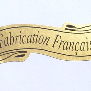 Può includere: Un'etichetta color oro con la scritta "Fabrication Française" in elegante corsivo. L'etichetta ha la forma di un banner con volute decorative. Il testo è in un classico carattere serif.