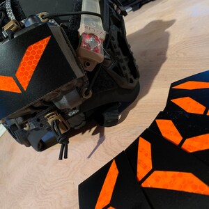 May include: Five black and orange reflective stickers with a chevron design. The stickers are arranged in a stack with the orange chevron facing up. The stickers are designed for use on helmets or other gear.