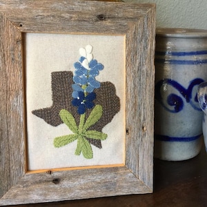 May include: A framed embroidery of a bluebonnet flower in the shape of the state of Texas. The frame is made of rustic wood.