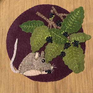 May include: A handcrafted textile art piece featuring a gray mouse with a burlap body, a sprig of green leaves, and dark berries on a purple background. The leaves have green stitching, and the berries are black beads.