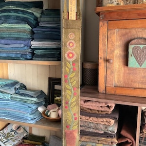 Puede incluir: Un porta agujas de tela marrón con un diseño de vid verde y bayas rojas. El porta agujas tiene una maceta roja en la parte inferior y está colgado de un estante de madera. El texto "FACTORY PAINT & DECOR" es visible en una regla de madera que cuelga de la parte superior del porta agujas.