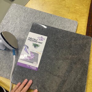 May include: Two gray felted wool pressing mats, one with a label that reads "13.5" SQUARE". A small white and blue iron is on the top left corner. The mats are on a light brown surface.