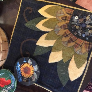 May include: A handmade quilted sunflower wall hanging with a denim background. The sunflower is made of felt and has a center made of buttons. The wall hanging is framed with a brown and gold trim.