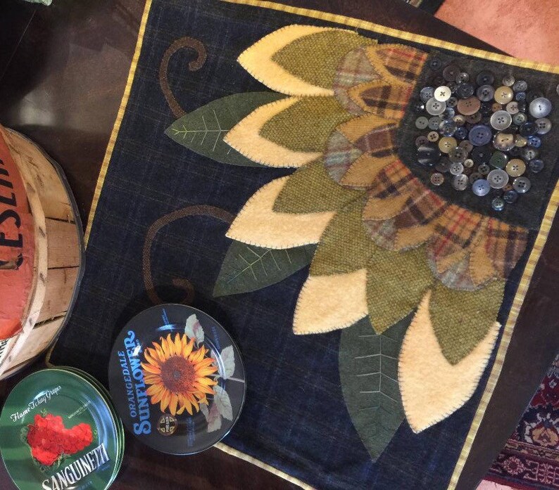 May include: A handmade quilted sunflower wall hanging with a denim background. The sunflower is made of felt and has a center made of buttons. The wall hanging is framed with a brown and gold trim.