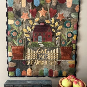 Puede incluir: Un tapiz de pared acolchado con un esquema de color marrón, rojo y verde. Presenta una pequeña casa roja con una puerta blanca y dos plantas en maceta con flores. El texto "Give me the simple life" está bordado en la parte inferior del tapiz.