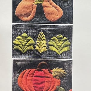 May include: A textile wall hanging featuring three stacked rectangular panels with dark gray backgrounds and intricate quilting. The top panel shows two orange pears, the middle shows three green leaves, and the bottom shows a pumpkin. Triple R Mercantile.