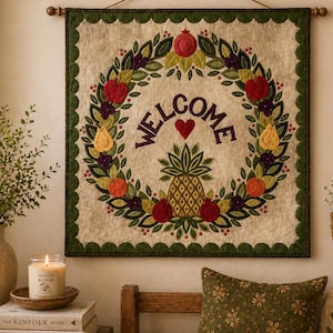 May include: A square wall hanging with a cream-colored background features a wreath design with the word "WELCOME" in purple. The wreath includes a pineapple, apples, pears, and pomegranates. The border is green with a scalloped edge. A wooden rod and string are used for hanging.