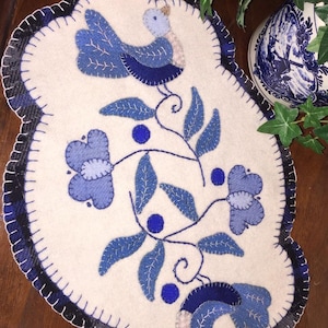 Puede incluir: Un camino de mesa de fieltro blanco con un dise&ntilde;o floral azul y blanco y dos p&aacute;jaros azules. El camino de mesa tiene un borde festoneado.