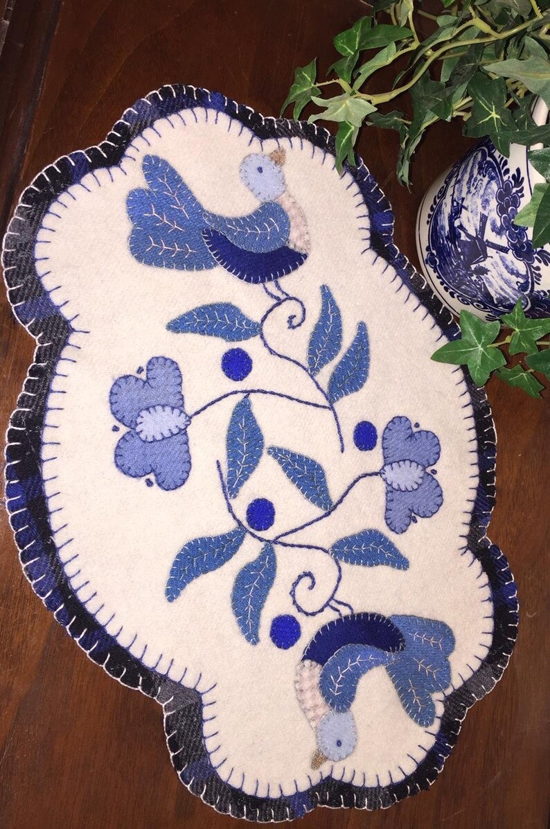 Puede incluir: Un camino de mesa de fieltro blanco con un dise&ntilde;o floral azul y blanco y dos p&aacute;jaros azules. El camino de mesa tiene un borde festoneado.