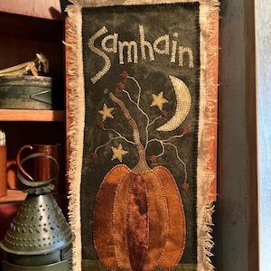 May include: A handmade wall hanging with a green background featuring a pumpkin, a crescent moon, and stars. The word "Samhain" is stitched in white thread at the top of the hanging.