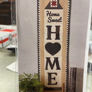 May include: A quilted wall hanging with a brown and beige background. The hanging features a house with a red star, the words "Home Sweet" and the word "HOME" in large black letters. The letters are outlined in white and the "O" is a heart shape.