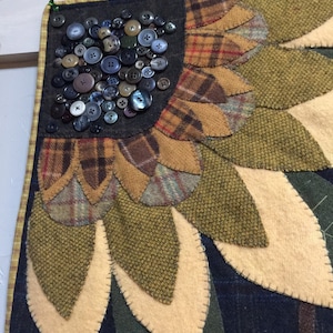 May include: A close-up of a handmade wool quilt depicting a sunflower with a center made of buttons. The sunflower is made of various shades of brown, green, and yellow wool fabric.