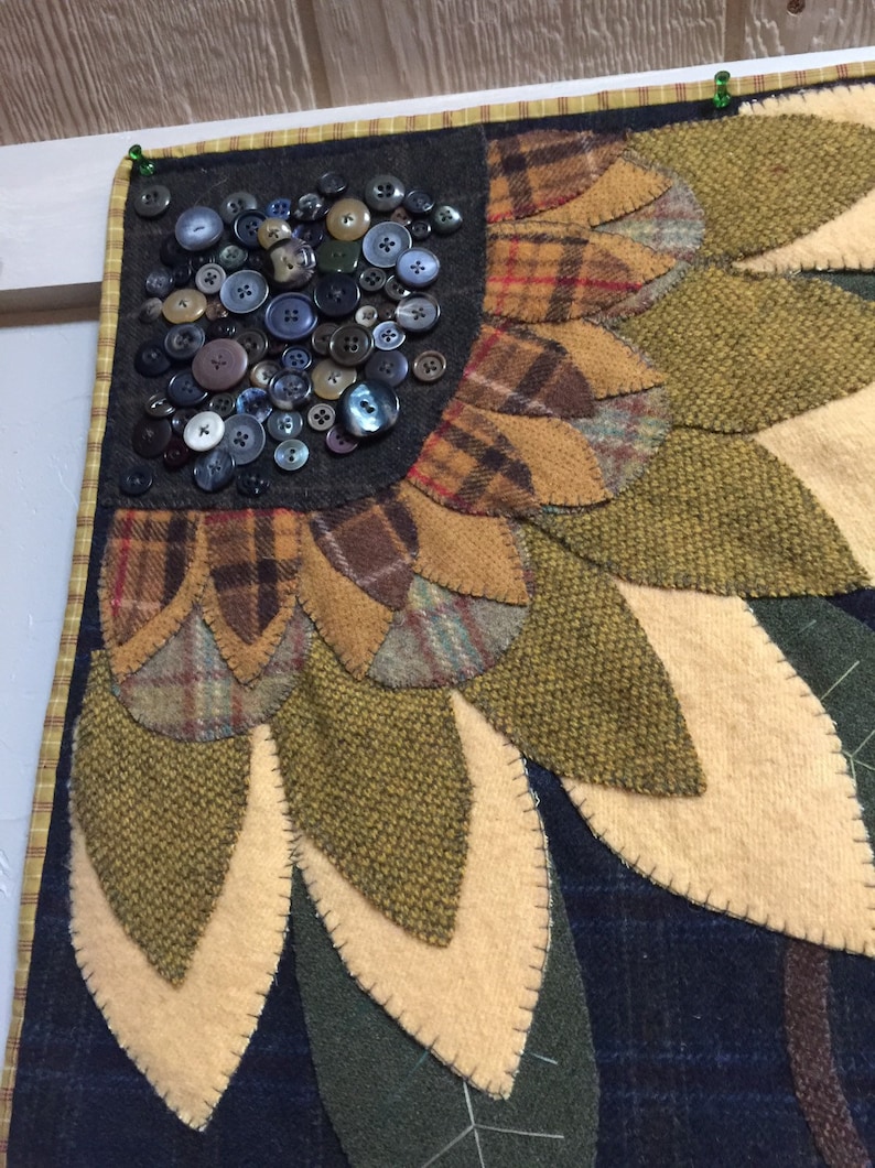 May include: A close-up of a handmade wool quilt depicting a sunflower with a center made of buttons. The sunflower is made of various shades of brown, green, and yellow wool fabric.
