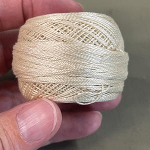 Puede incluir: Una bola de hilo blanco roto, probablemente hilo de bordar o ganchillo. El hilo es liso y parece estar hecho de una fibra natural. La bola se sostiene en una mano, mostrando la escala del hilo. Ideal para proyectos de manualidades.