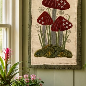 May include: A fabric wall hanging depicting three red mushrooms with white spots, grey stems, and green grass. The artwork is framed with a green border and hangs from a wooden dowel. The background is a light beige.