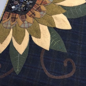 May include: A handmade wool felt wall hanging featuring a sunflower with brown, green, and yellow petals. The sunflower is surrounded by green leaves and a brown swirl design on a dark blue background.
