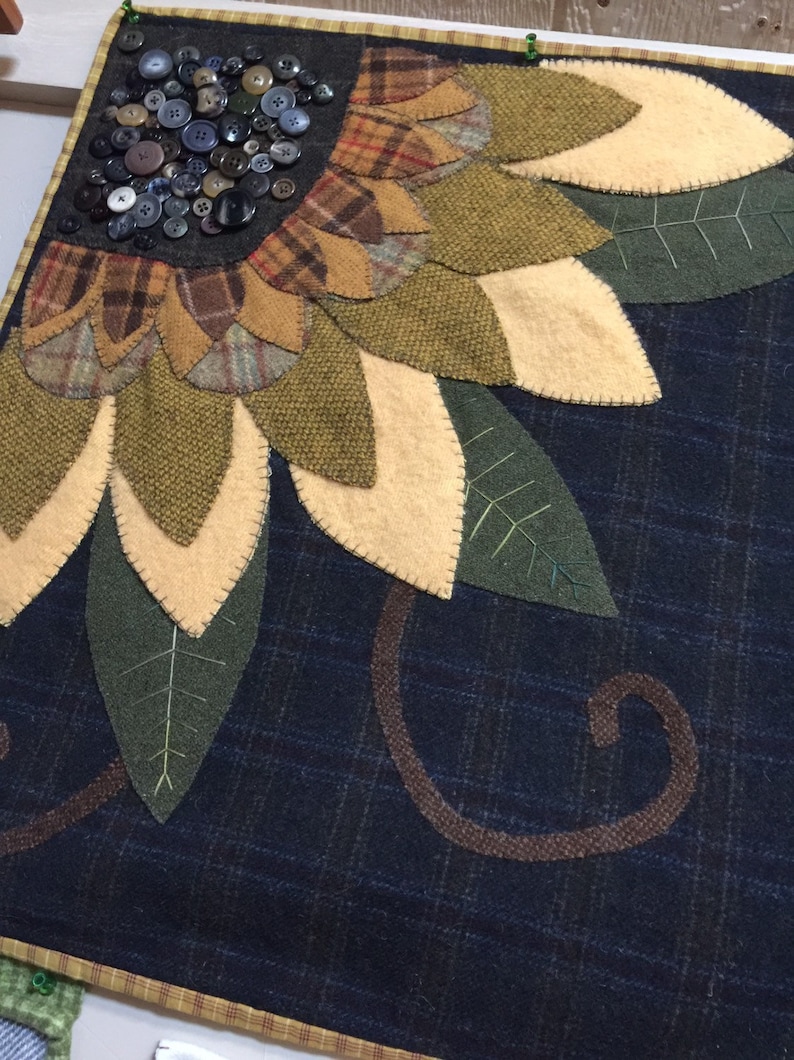 May include: A handmade wool felt wall hanging featuring a sunflower with brown, green, and yellow petals. The sunflower is surrounded by green leaves and a brown swirl design on a dark blue background.