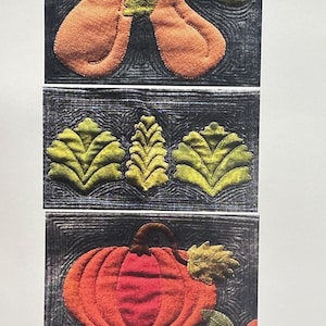 May include: A textile wall hanging featuring three stacked rectangular panels with dark gray backgrounds and intricate quilting. The top panel shows two orange pears, the middle shows three green leaves, and the bottom shows a pumpkin. Triple R Mercantile.