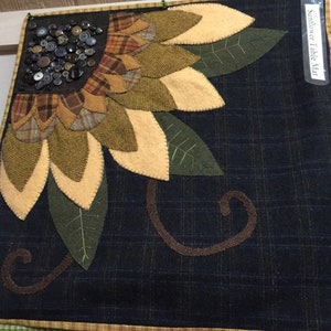 May include: A handmade sunflower table mat with a dark blue background and a yellow, green, and brown sunflower design. The sunflower is made of felt and has a button center. The mat has a brown and yellow border.