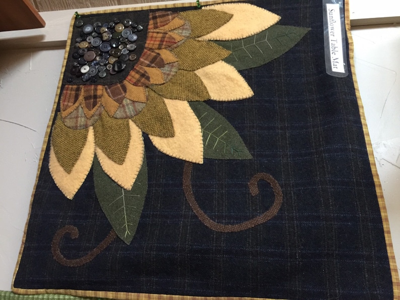 May include: A handmade sunflower table mat with a dark blue background and a yellow, green, and brown sunflower design. The sunflower is made of felt and has a button center. The mat has a brown and yellow border.
