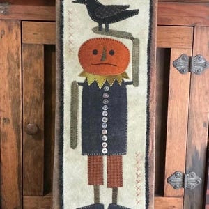 Może przedstawiać: Ozdoba ścienna w stylu folk art z postacią z dyniową głową i czarnym krukiem. Postać ma ciemnoniebieski płaszcz z guzikami, brązowe spodnie i czarne buty. Tło jest kremowe z dekoracyjnymi przeszyciami.