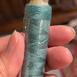 以下が含まれることがあります： 「Wool EN07」とラベルが付いた、ティールグリーンの刺繍糸の糸巻きです。