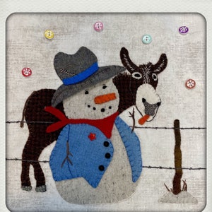 May include: A felt snowman wearing a blue jacket, red scarf, and a cowboy hat is standing next to a brown donkey. The donkey is holding a stick in its mouth. The background is a light gray fabric with colorful buttons scattered around.