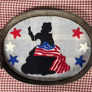 Puede incluir: Una bandeja decorativa de metal con un diseño de fieltro que presenta una silueta negra de una mujer con una bandera estadounidense drapeada. El diseño incluye estrellas rojas, blancas y azules. La bandeja tiene dos asas y está sobre un fondo a cuadros rojos y blancos.