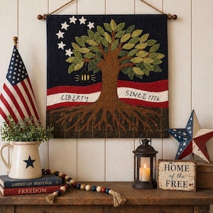 May include: A patriotic wall hanging with a tree design, stars, and the words "LIBERTY" and "SINCE 1776". The image also features an American flag, a star-shaped decoration, and a candle lantern, all arranged on a wooden table.